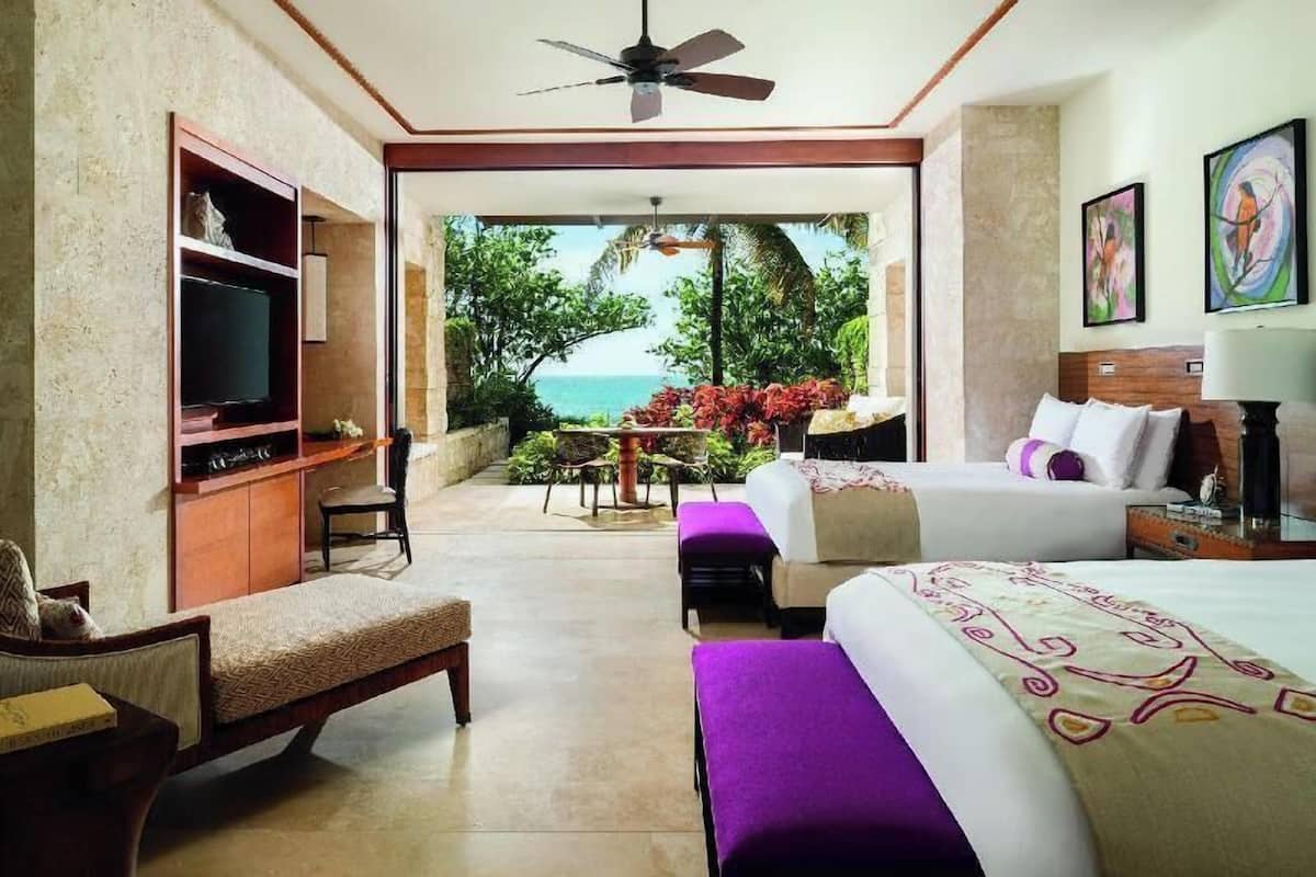 Habitación de hotel con cama, televisor, zona de estar con mesa y sillas, y vista al océano.