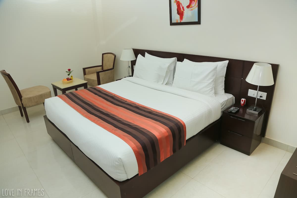 A hotel room with a large bed, a bedside table with a lamp, a chair, and a small table with fruit and a flower.