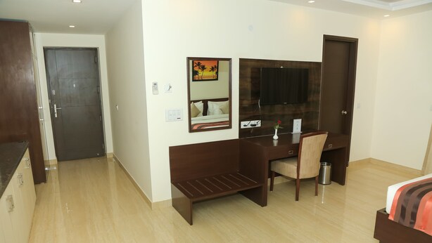 A hotel room with a bed, a desk, a chair, a small table with a vase and fruit, a mirror, and a TV. - Treehouse Neo Casa-Unit of Neo Residency (Gurugram)