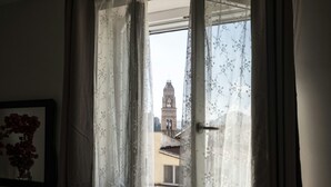 A room with a window featuring floral curtains, a framed picture of flowers, and a view of a building with a tower. - Santa Lucia B&B (Gaeta)