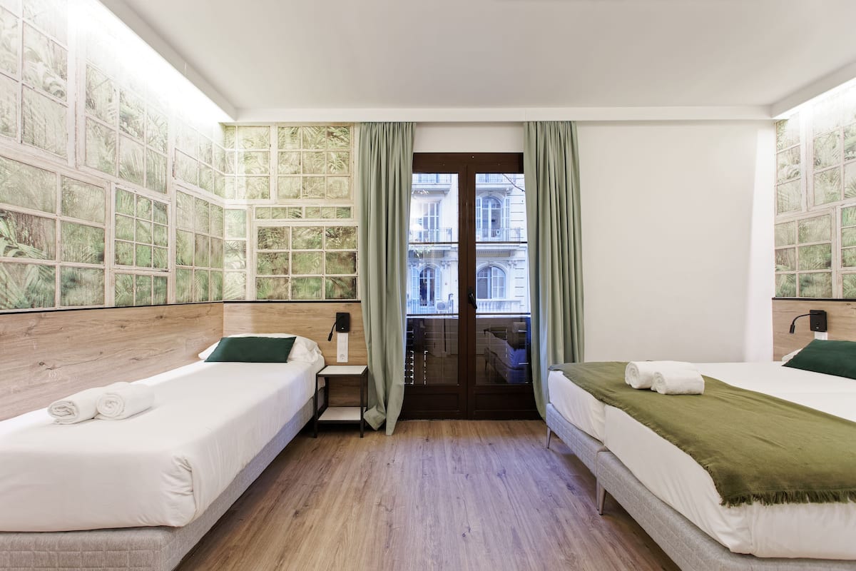 A hotel room with two beds, a wooden headboard, a small bedside table, a window with green curtains, and a view of a building through the window.