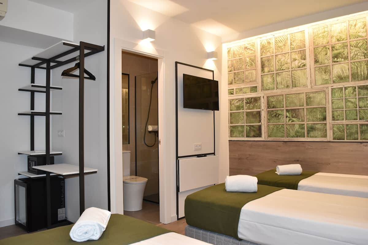 A hotel room with two beds, a TV, a shower, and a window with a view of greenery.