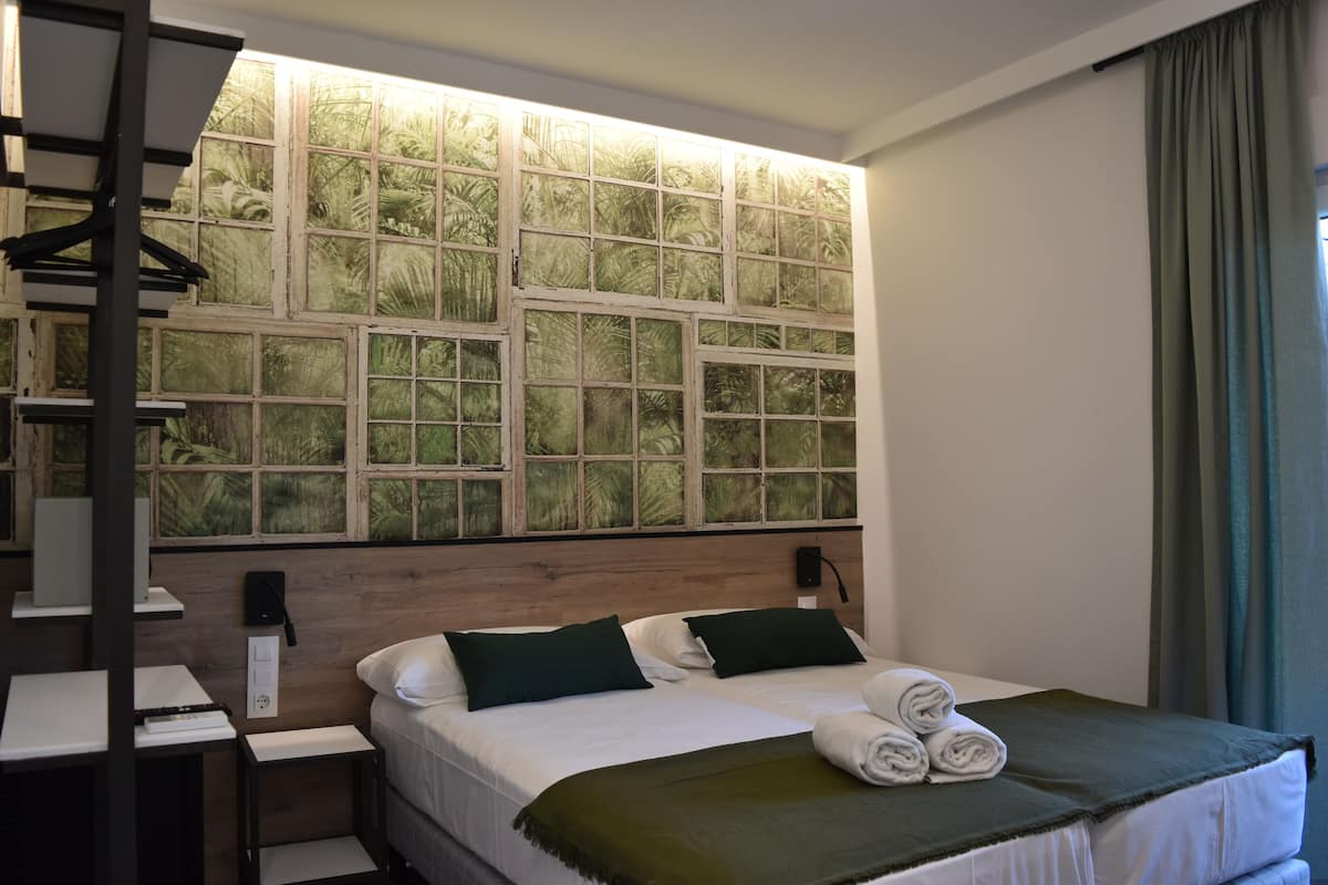 A hotel room with a large window, a bed with green pillows, a bedside table, and a wall-mounted TV.