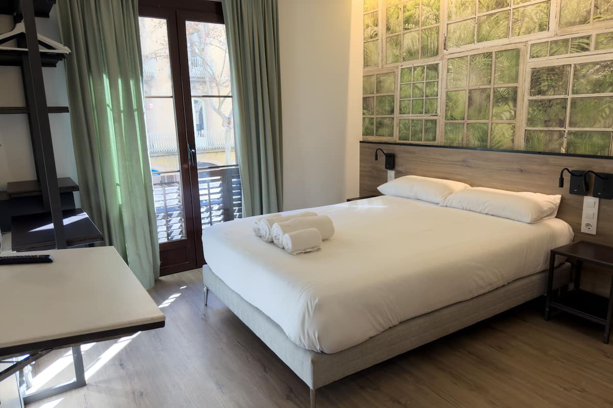 A hotel room with a bed, a desk, a window with curtains, and a wall with a green leafy pattern.