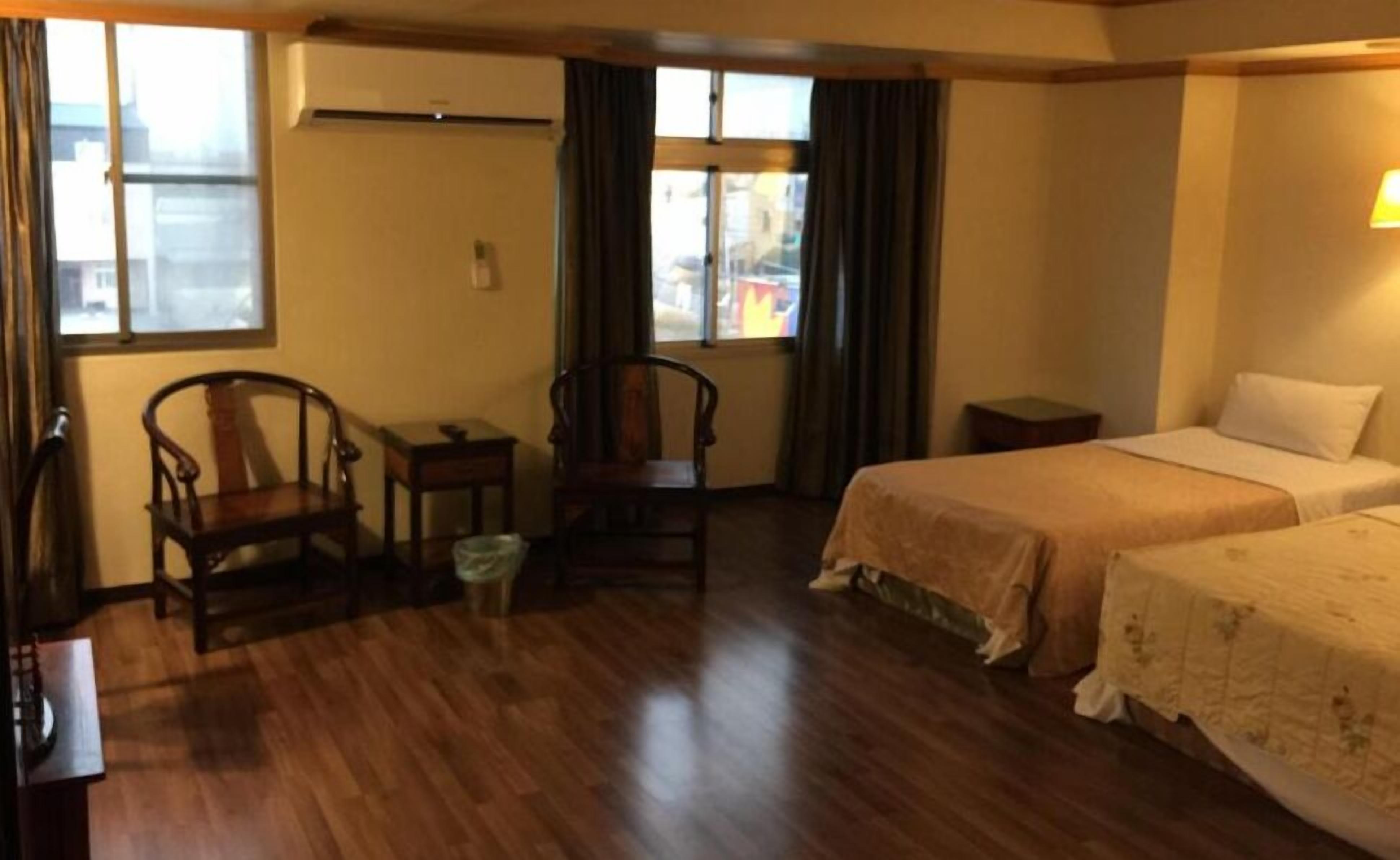 A hotel room with a wooden floor, two single beds, a chair, a small table, and a window with curtains.