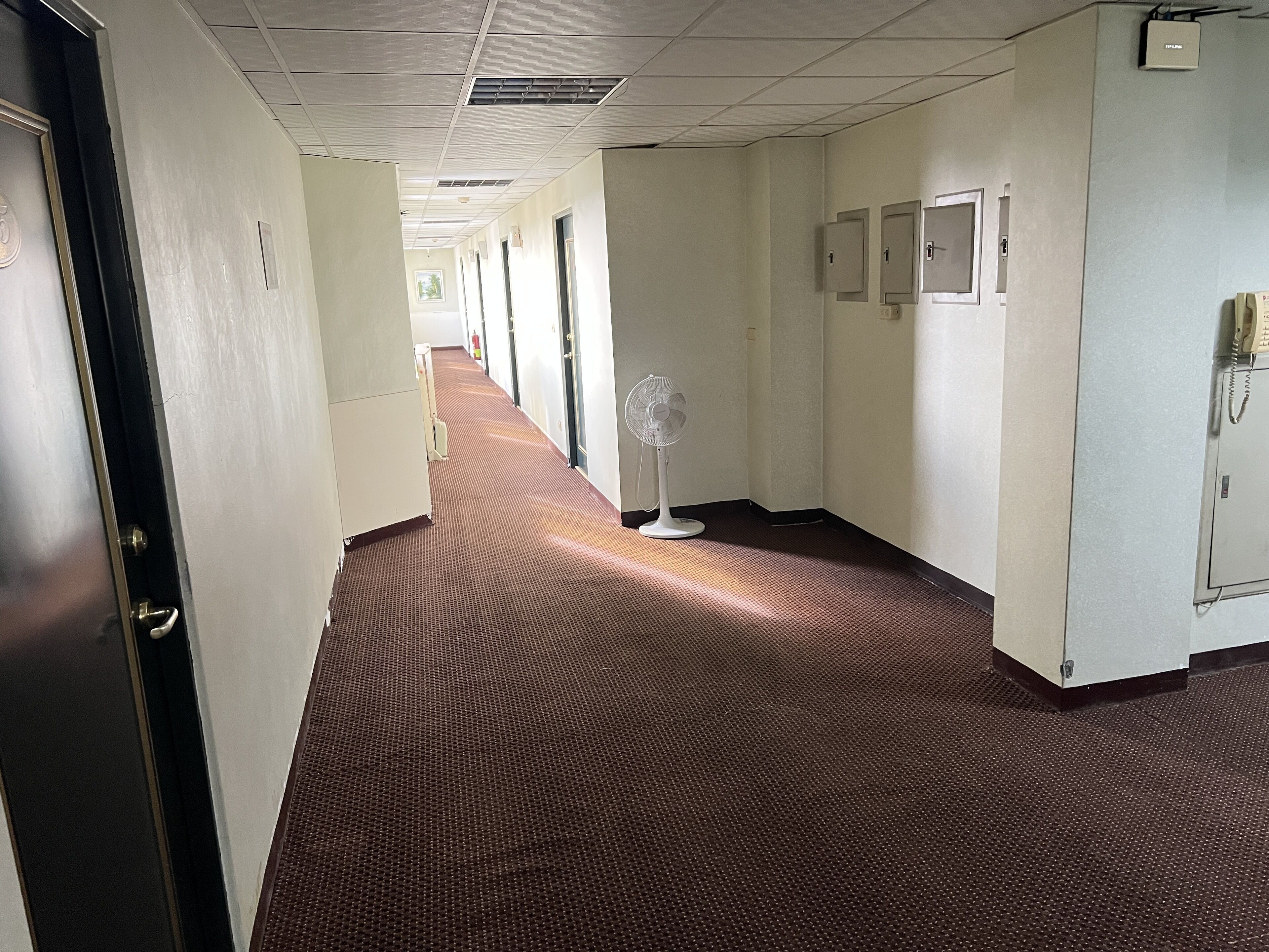 Un pasillo de hotel con suelo alfombrado, puertas y un ventilador.