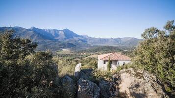 Un pequeño edificio de techo rojo, rodeado de terreno pedregoso y vegetación densa, con vistas a un valle y montañas a lo lejos.