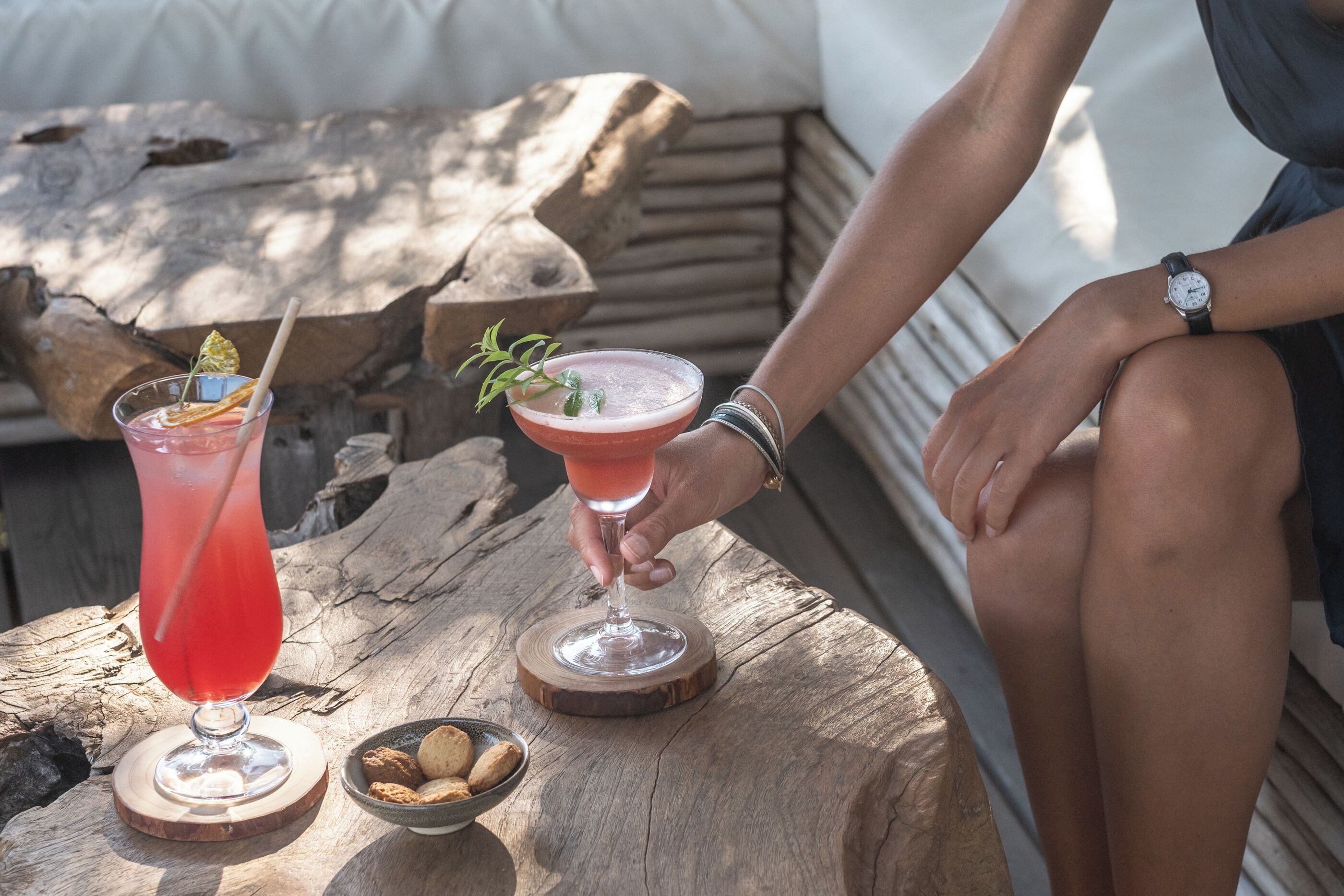 Una mesa de madera con dos bebidas, una en un vaso alto y otra en una copa de martini, y un tazón pequeño de galletas.