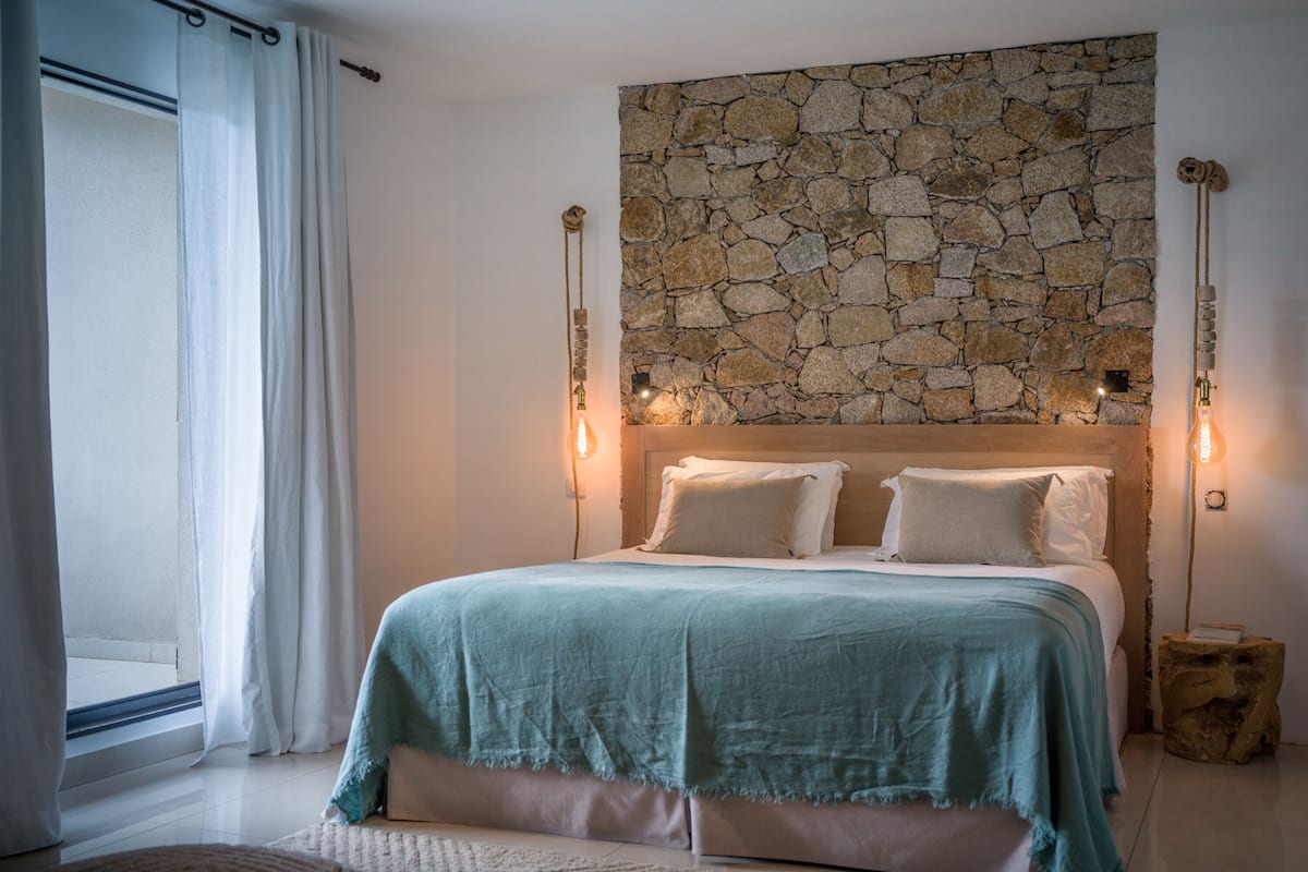 A bedroom with a stone wall, a bed with white linens, and two wall-mounted light fixtures.