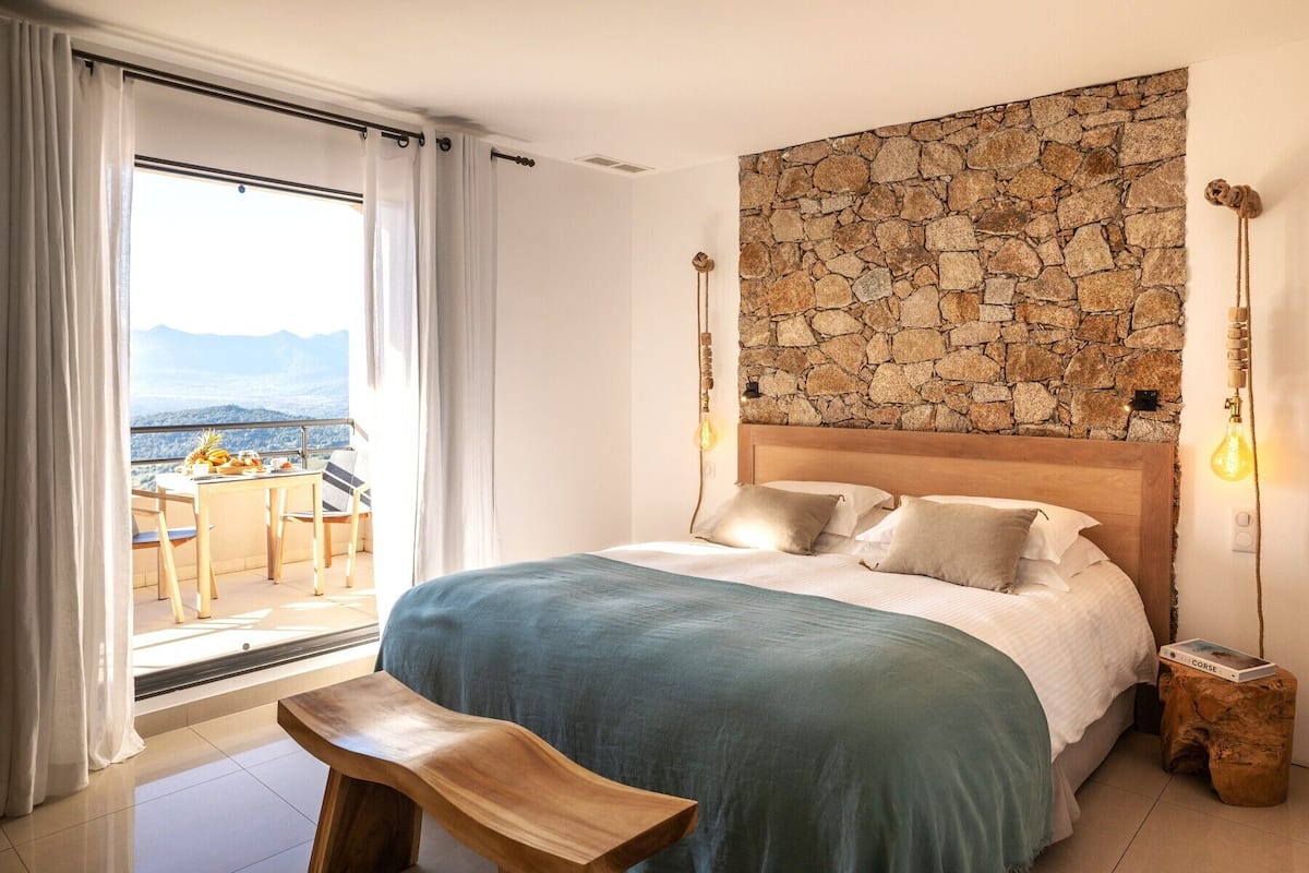 A bedroom with a stone wall, a bed with pillows, a bench, and a view of mountains through a large window.