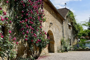A stone building with a covered courtyard, a weather vane, and a rose bush. - Secret Pyrénées (Lasseube)