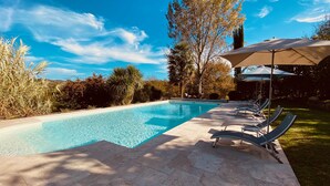A rectangular swimming pool with clear blue water, surrounded by a stone deck. There are lounge chairs with umbrellas and a stone wall in the background. - Secret Pyrénées (Lasseube)