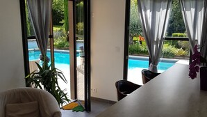 A modern living room with a sofa, a dining table, and a view of a swimming pool through large windows. - La Griotte Chambre D'hotes (Honfleur)