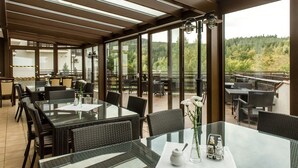 Un área de comedor al aire libre cubierta con mesas de vidrio y sillas de mimbre, con vistas a un paisaje boscoso.