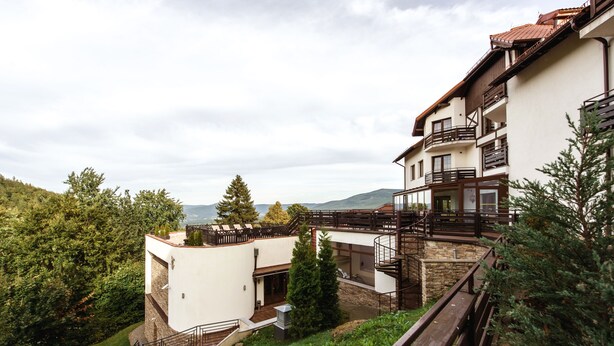 A multi-story building with balconies, a stone staircase, and a view of the surrounding greenery and hills.