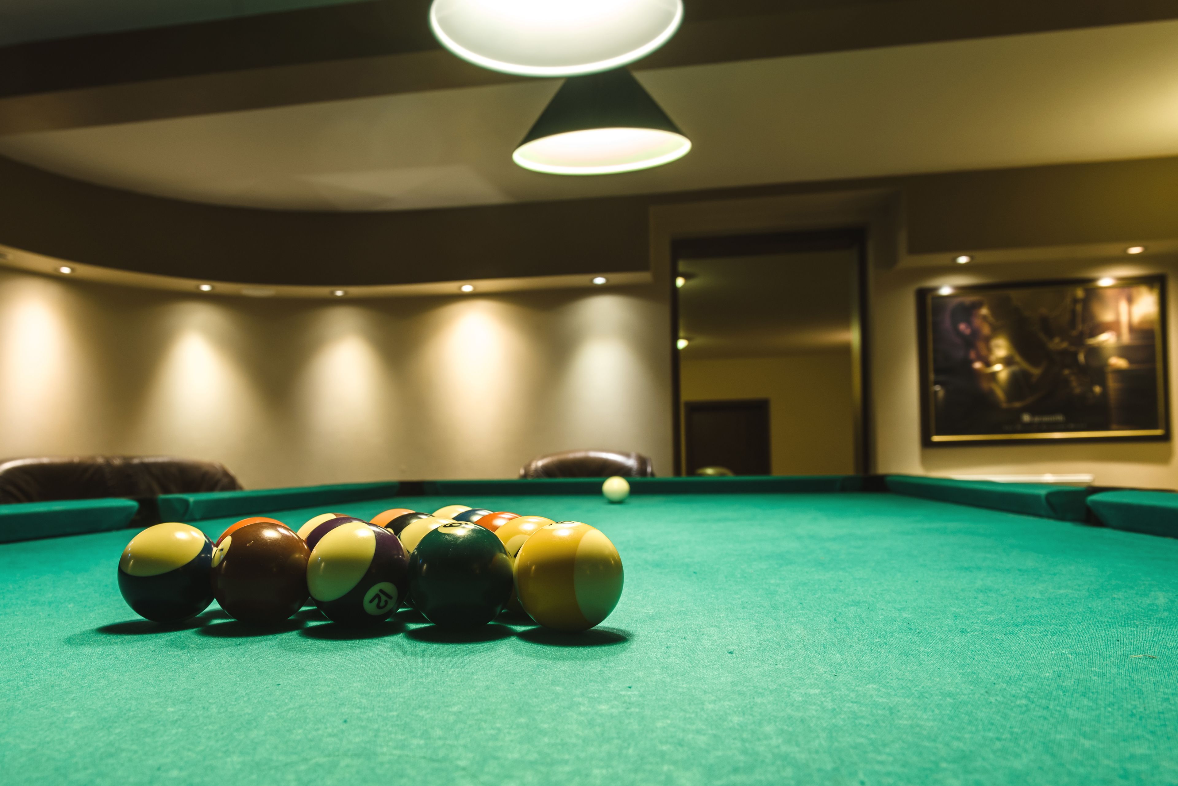 Une table de billard avec les billes alignées, un luminaire au plafond et un tableau encadré accroché au mur.