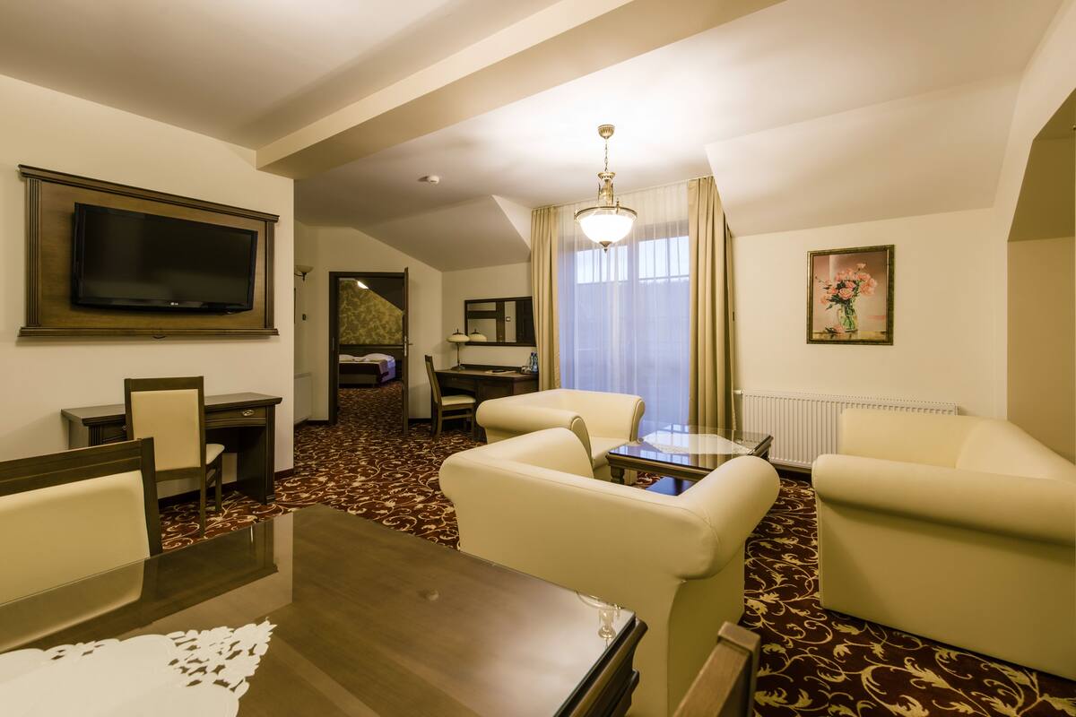 Habitación de hotel con televisor de pantalla plana, una mesa de comedor, dos sillones, una mesita, un candelabro, un cuadro y piso alfombrado.