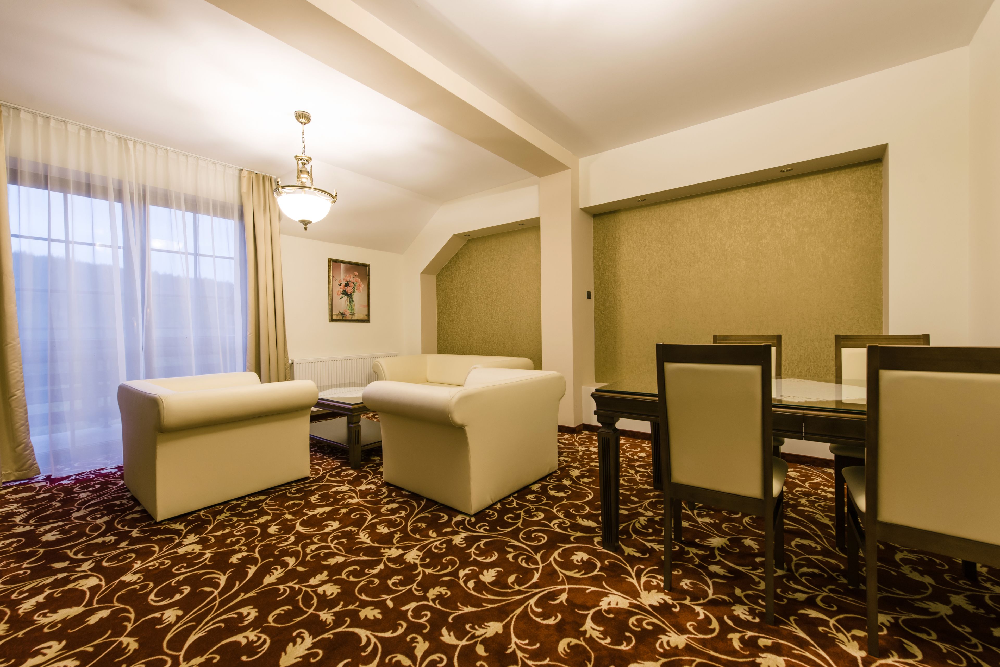 A hotel room with a brown patterned carpet, a sofa, a dining table with chairs, and a chandelier.