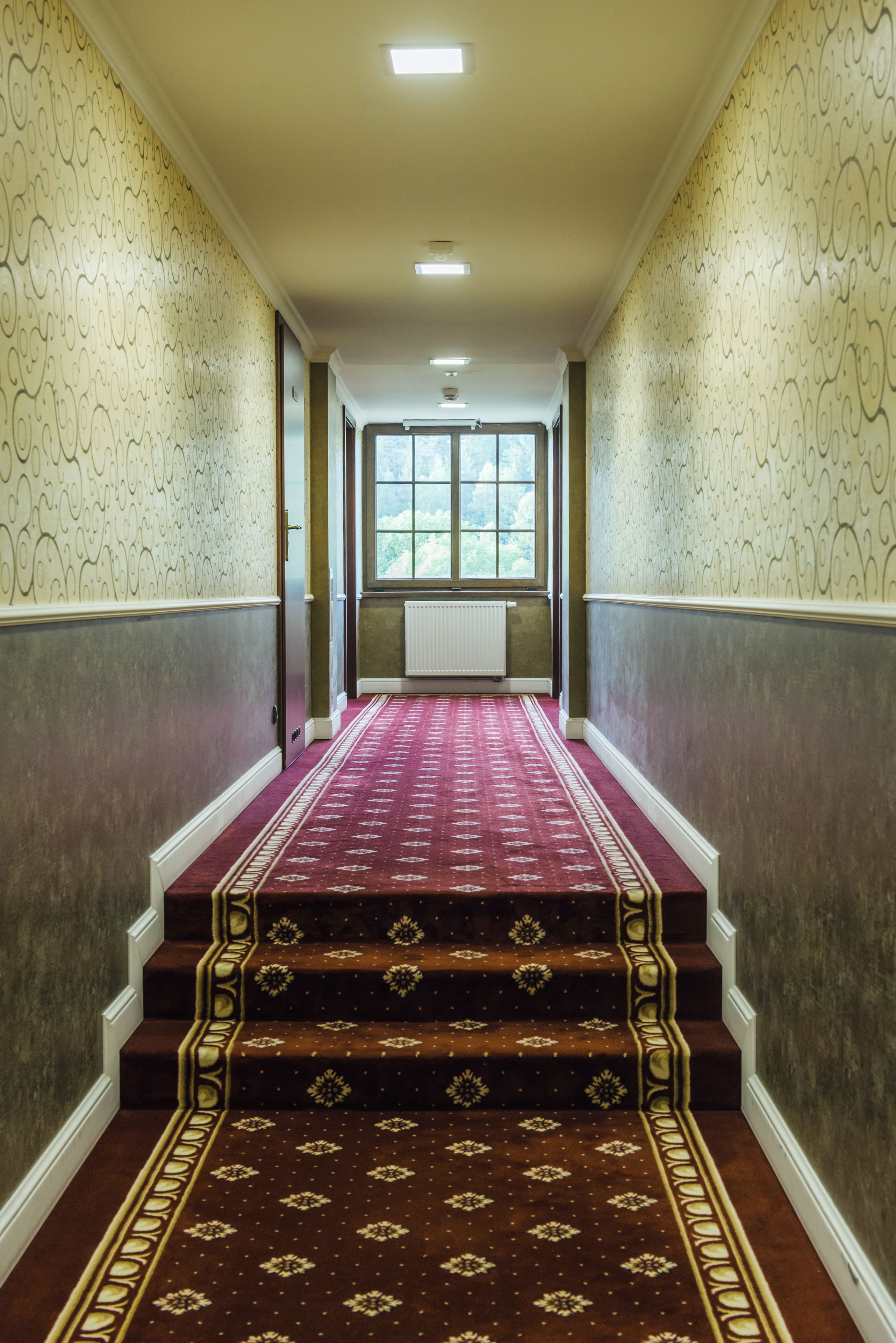 Un pasillo con papel tapiz estampado, alfombra roja y una escalera que lleva a una ventana.