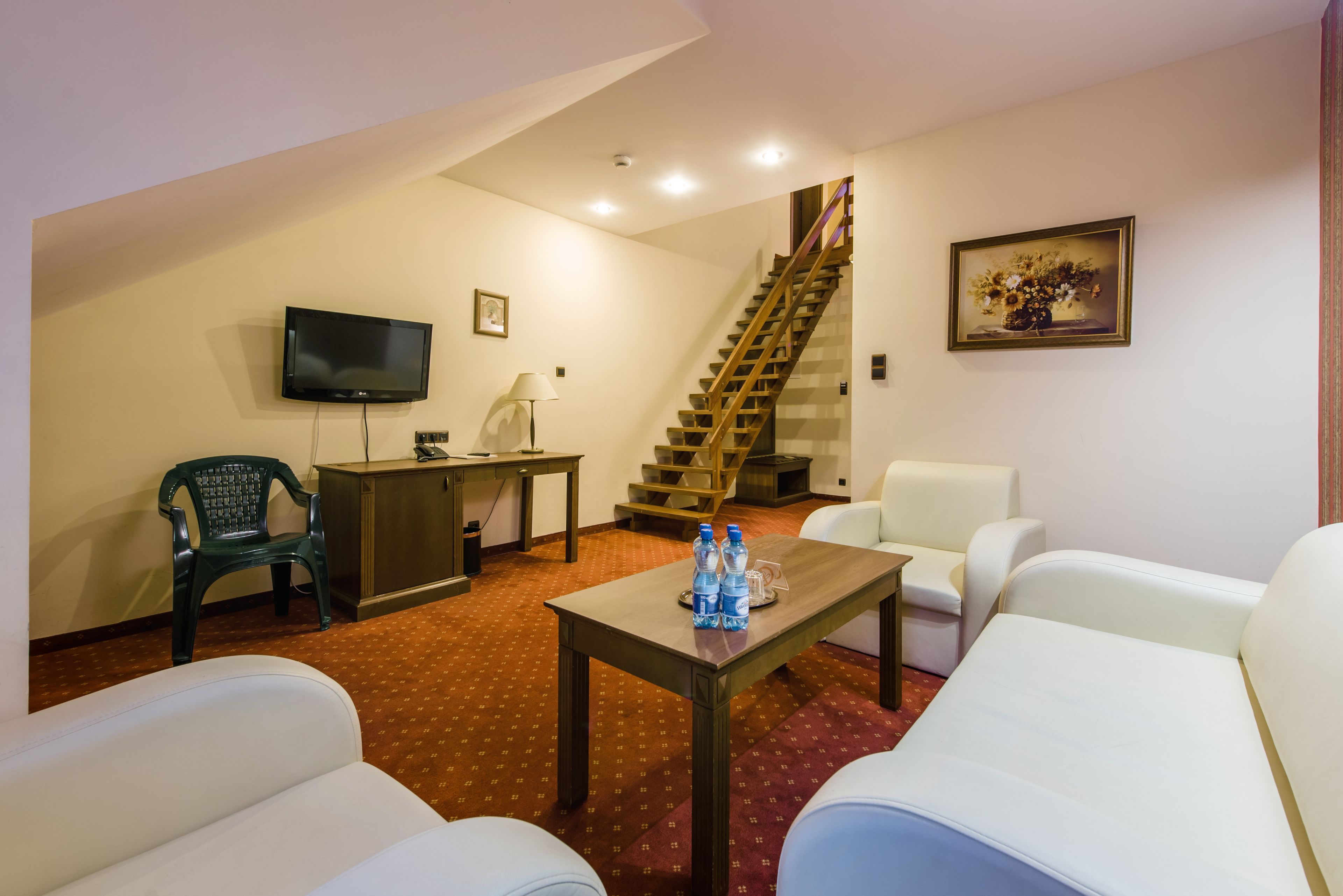A hotel room with a TV, a desk, a chair, a sofa, a staircase, and a painting on the wall.