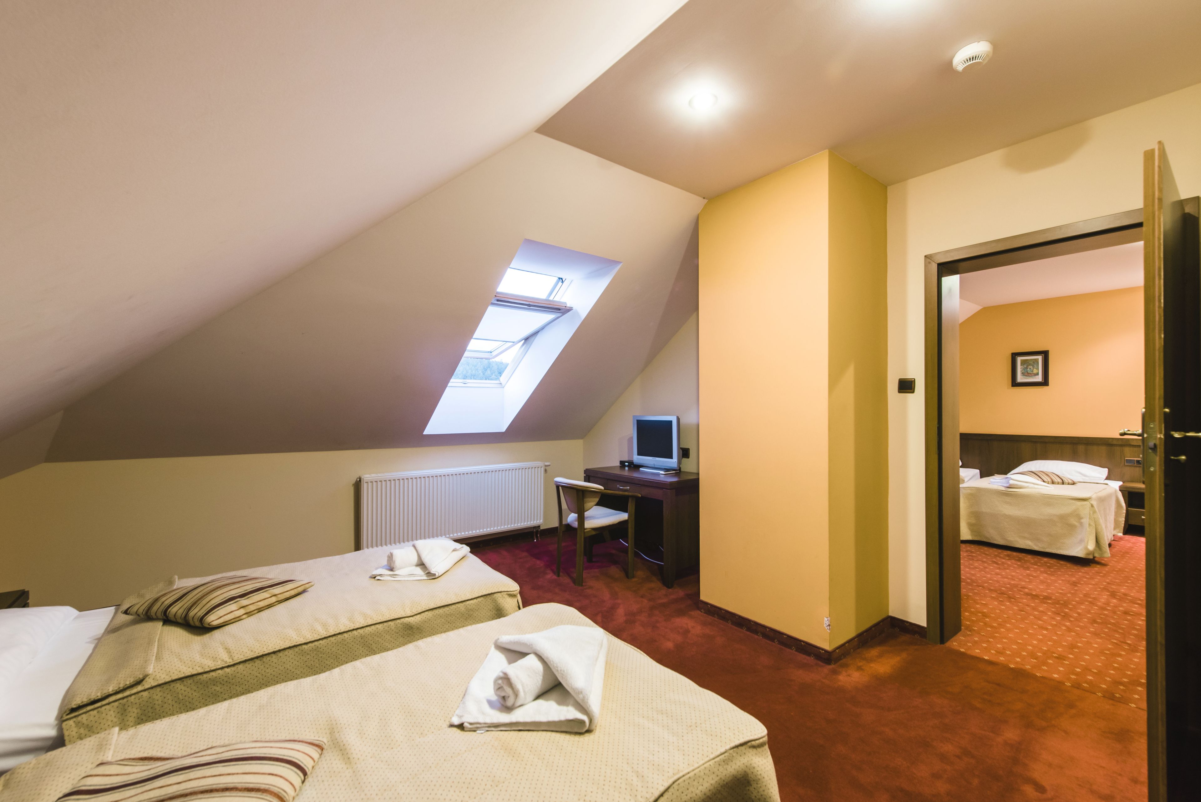A hotel room with two beds, a desk with a computer, a window, and a door leading to another room.