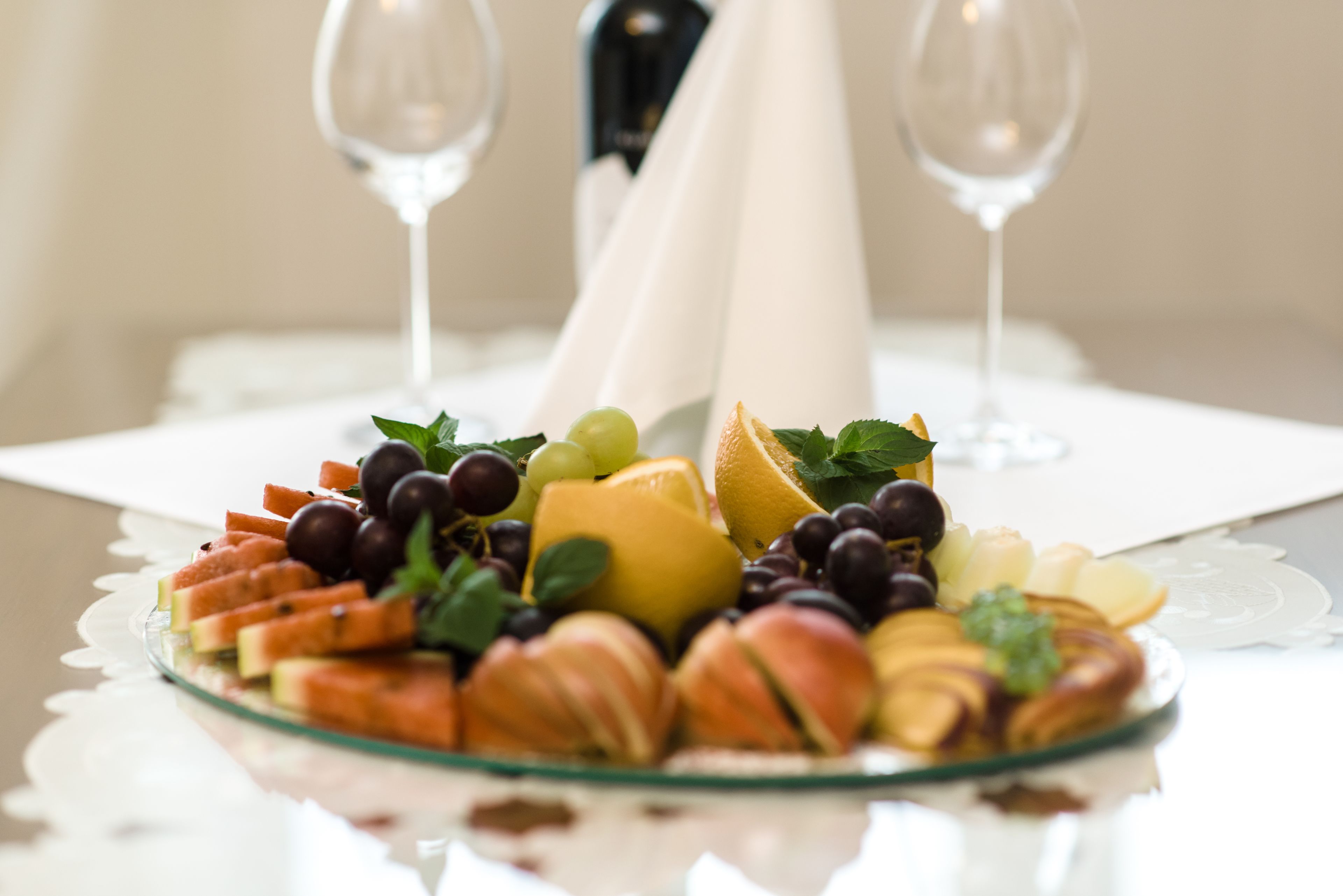 Een fruitschaal op een glazen bord met een servet en twee wijnglazen op de achtergrond.