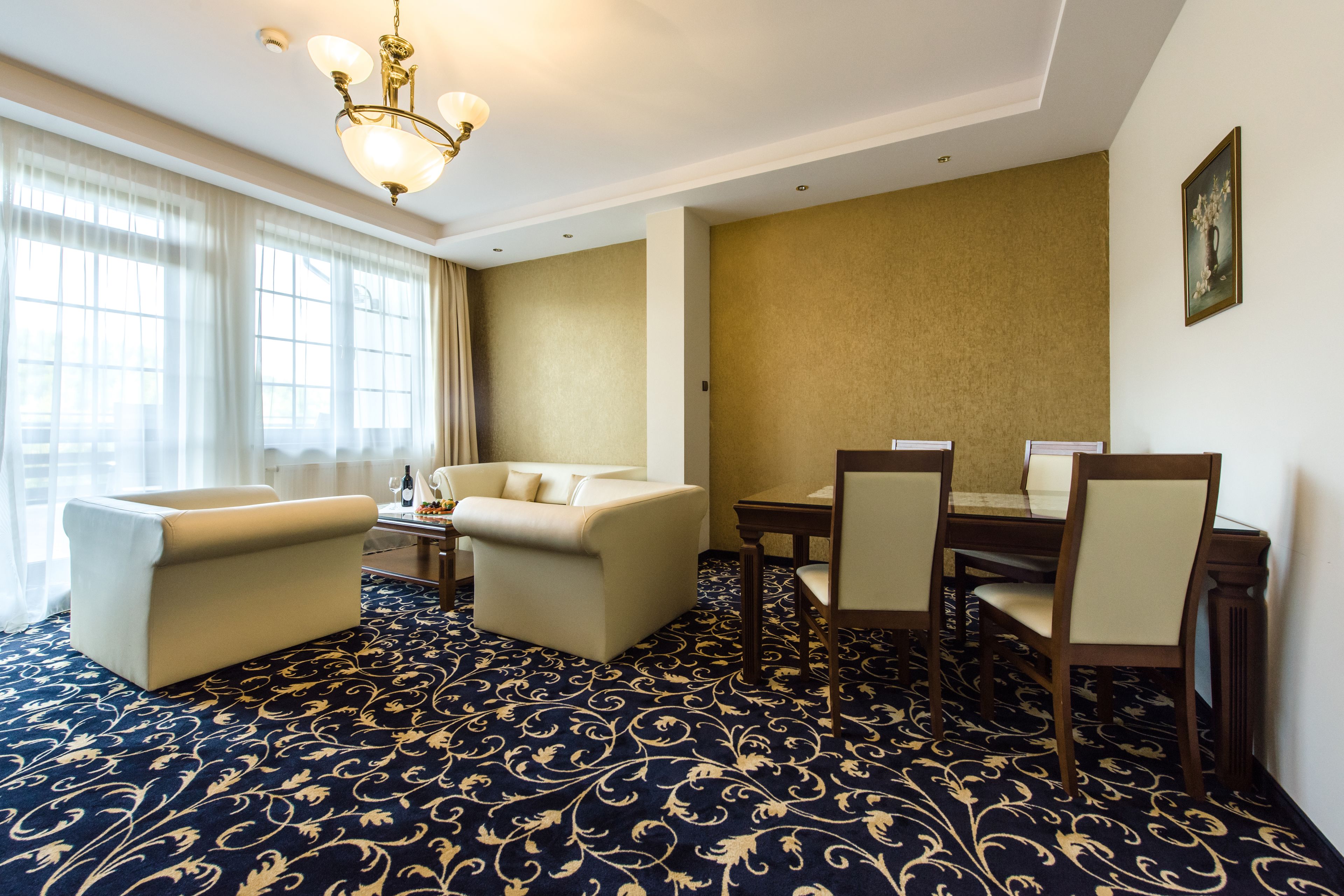 A hotel room with a large window, a sofa, a dining table with chairs, and a patterned carpet.