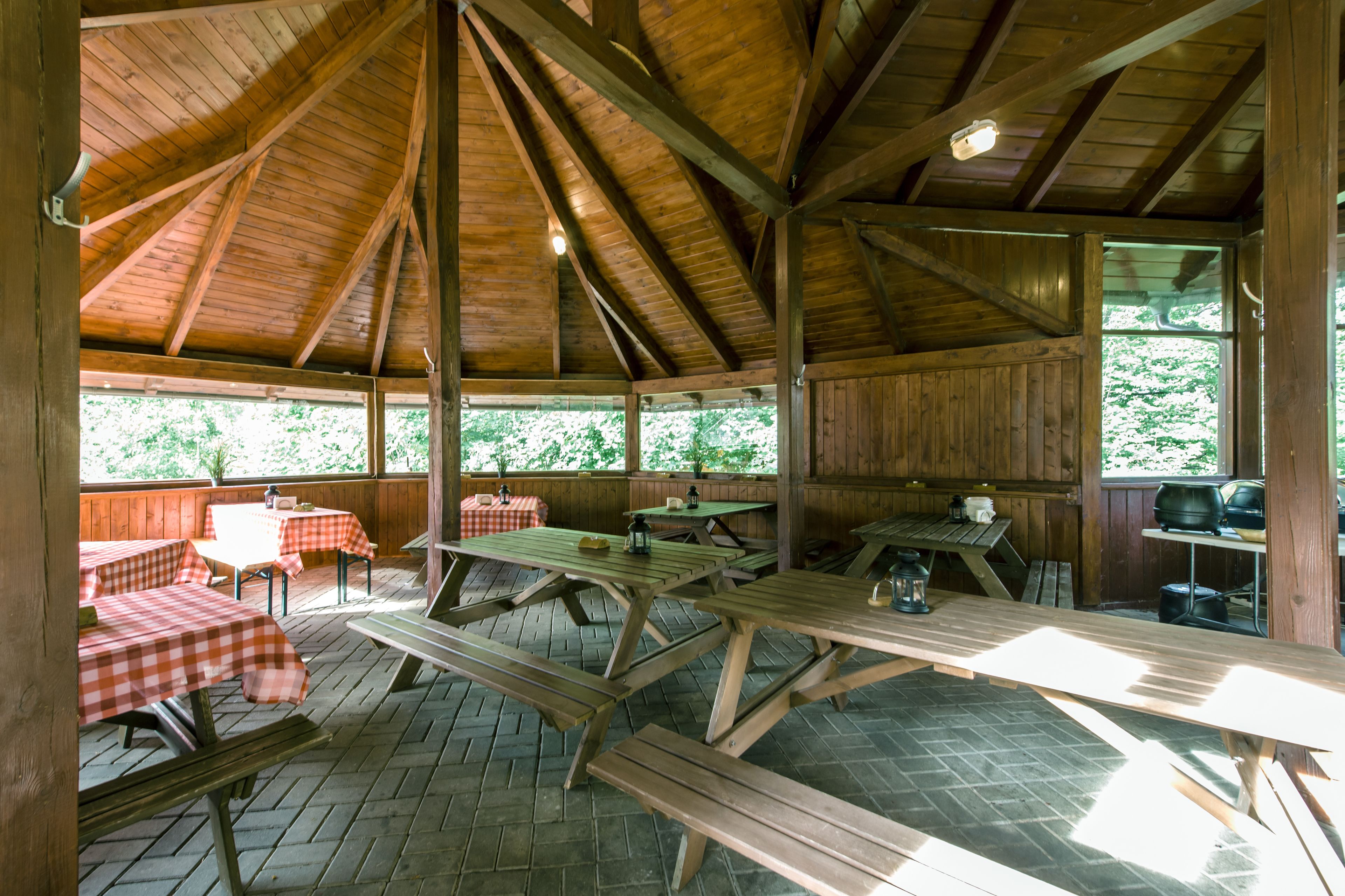 A covered picnic area with wooden benches and tables, some with checkered tablecloths, and a view of greenery outside.