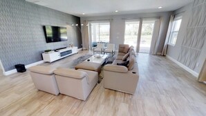 A spacious living room with a large sectional sofa, a flat-screen TV mounted on the wall, and a dining area with a table and chairs.