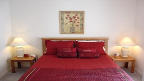 Uma cama bem arrumada com roupa de cama vermelha, duas mesas de cabeceira com abajures e uma obra de arte emoldurada acima da cama.