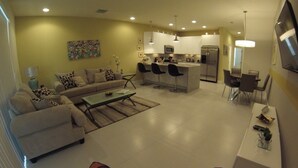 A modern kitchen with white cabinets, a central island with bar stools, and a dining area with a TV mounted on the wall. - Aco63664 - Serenity - 3 Bed 3 Baths Townhouse (Clermont)
