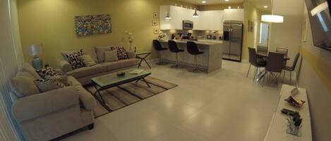 A modern kitchen with white cabinets, a central island with bar stools, and a dining area with a TV mounted on the wall.