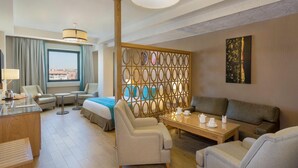 A hotel room with a bed, a sofa, a dining table, and a view of the city through a window. - Plus Hotel Setif (Setif)