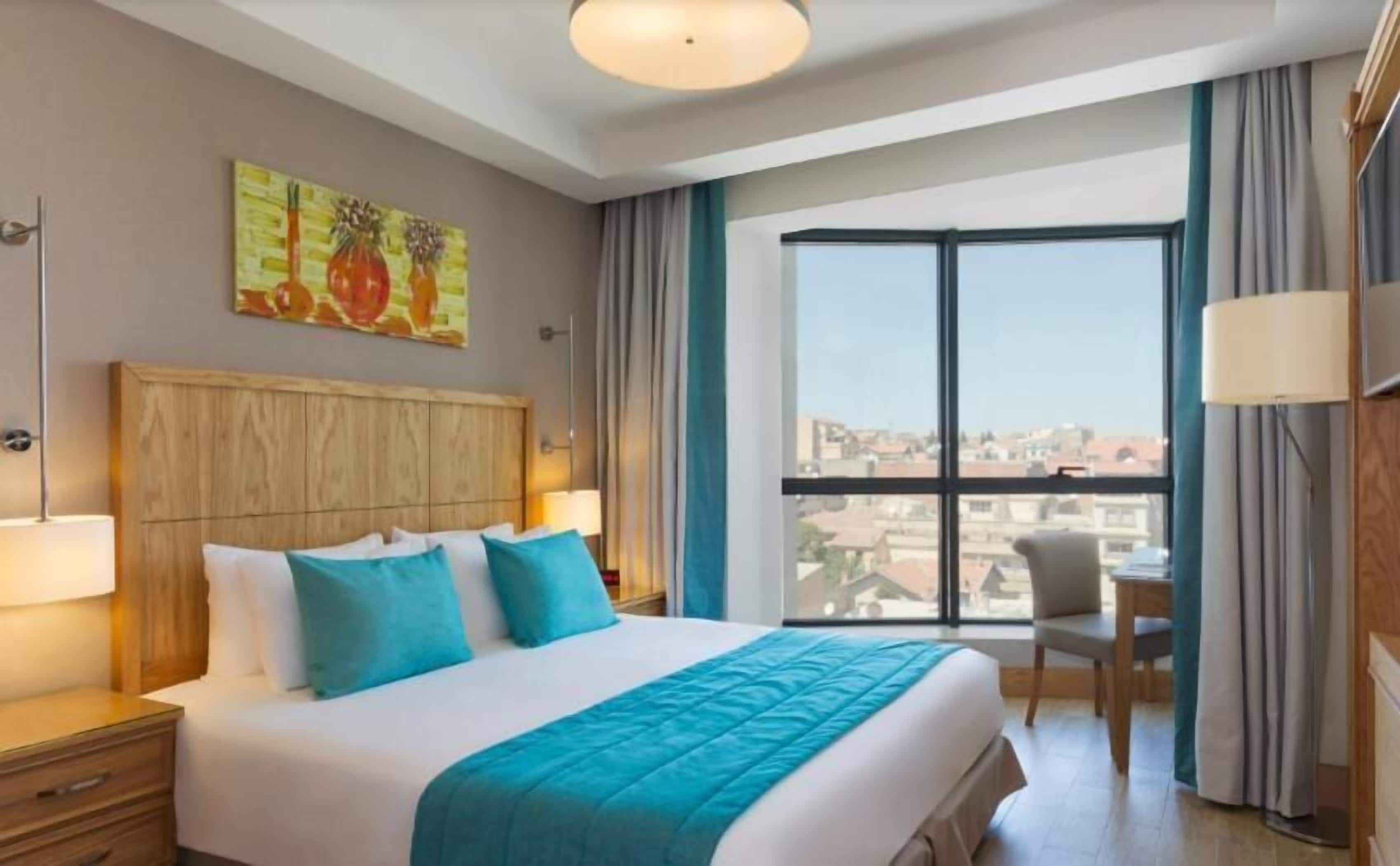 A hotel room with a large bed, a wooden headboard, a painting on the wall, and a view of the city through the window.