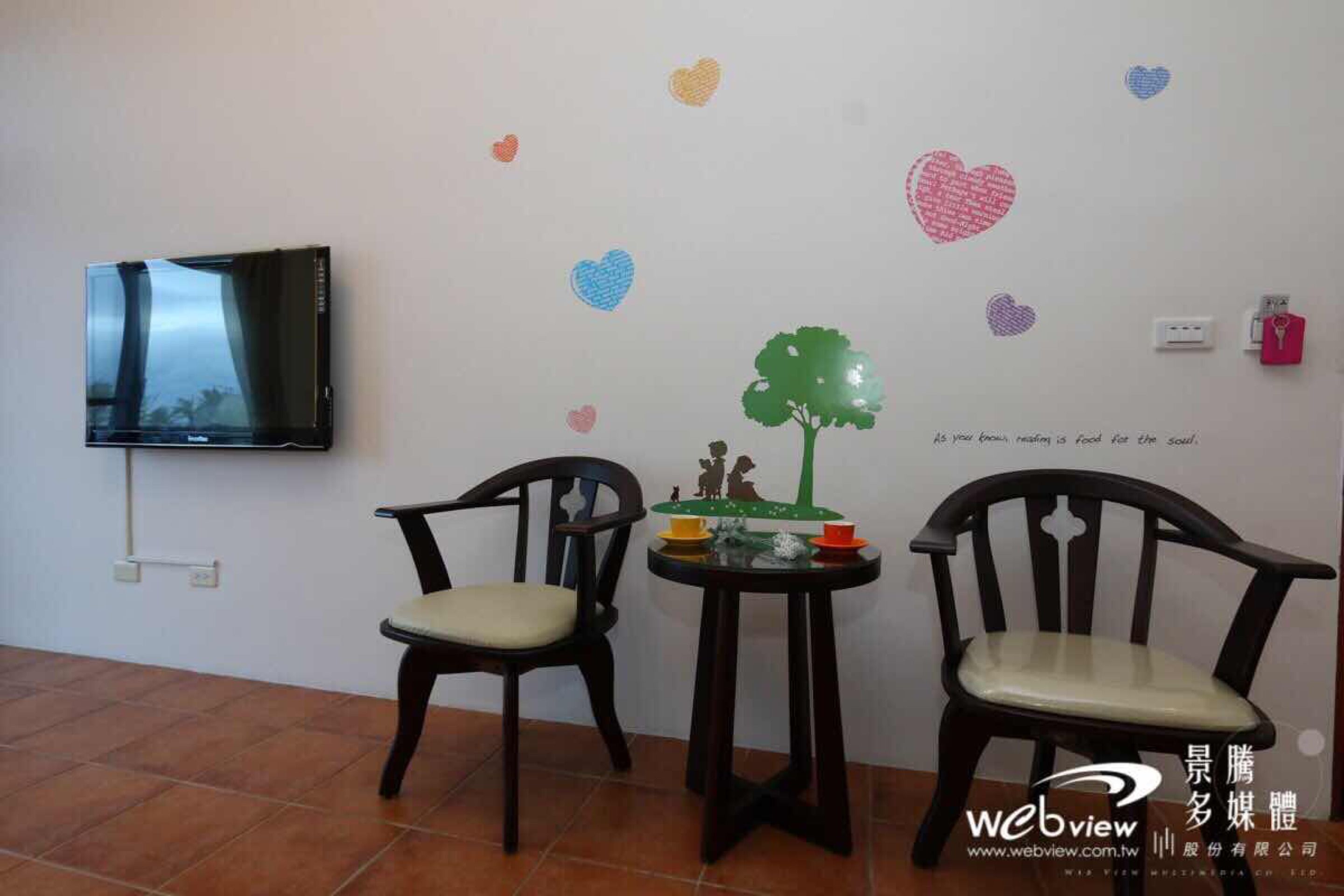 A room with a television mounted on the wall, two wooden chairs, a small table with a decorative tree and figurines, and heart-shaped wall decals.