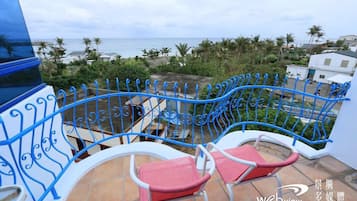 A balcony with blue metal railings, two red cushioned chairs, and a view of the sea and palm trees.