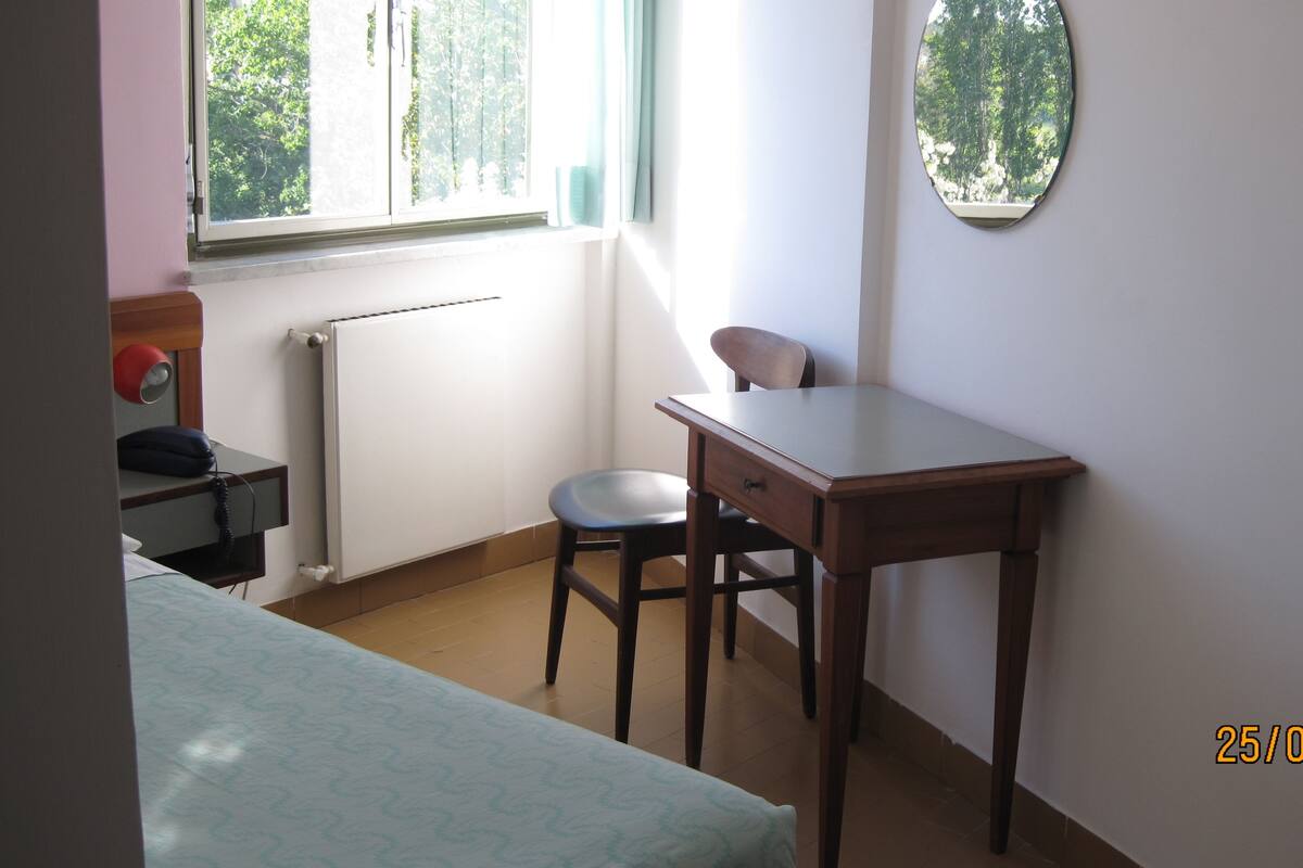 Una habitación pequeña y escasamente amoblada con una cama individual, un escritorio, una silla y un radiador.
