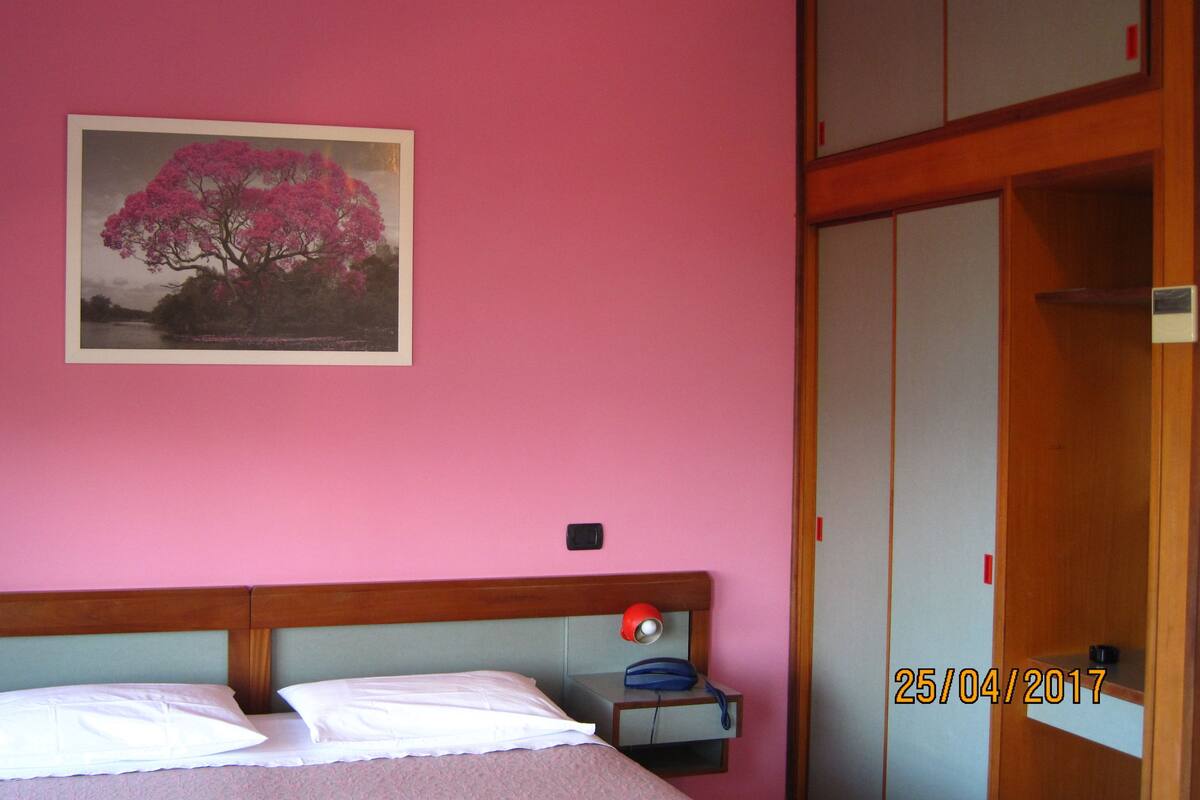 Un dormitorio con pared rosa, una cama con ropa blanca, una mesita de noche con lámpara y un cuadro con una imagen de un árbol.