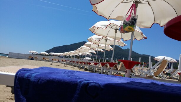 A beach with rows of white umbrellas and red chairs, a clear blue sky, and a mountain in the background. - Hotel Le Pleiadi (San Felice Circeo)
