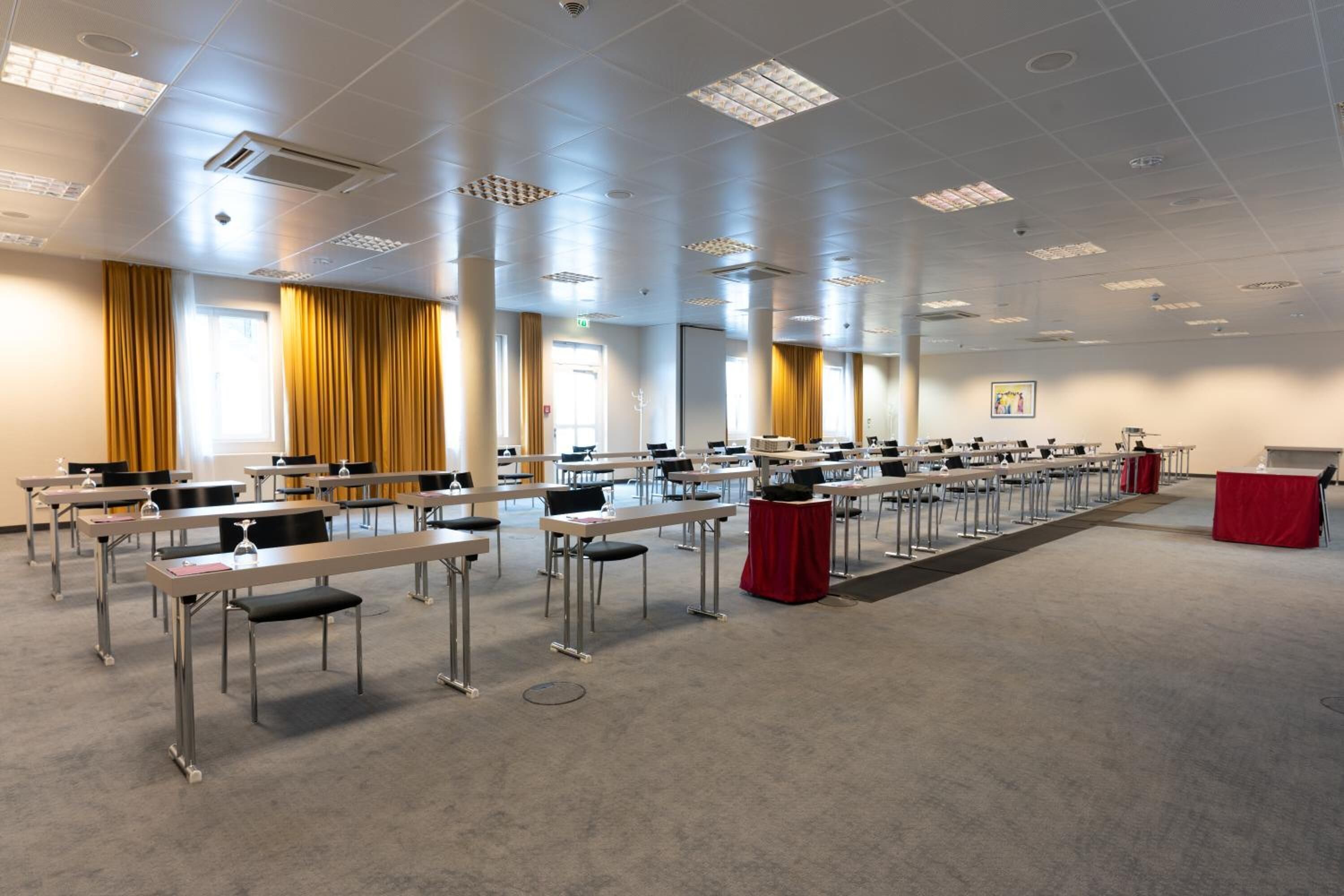 A conference room with rows of tables and chairs, a podium, and large windows with curtains.