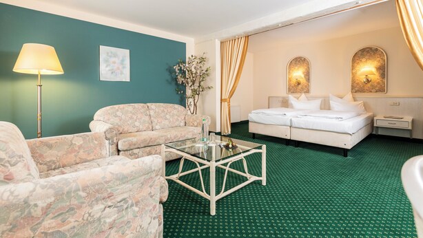Habitación de hotel con alfombra verde, sofá estampado con motivos florales, una mesita de centro de cristal, cama con ropa blanca y dos lámparas fijadas a la pared.