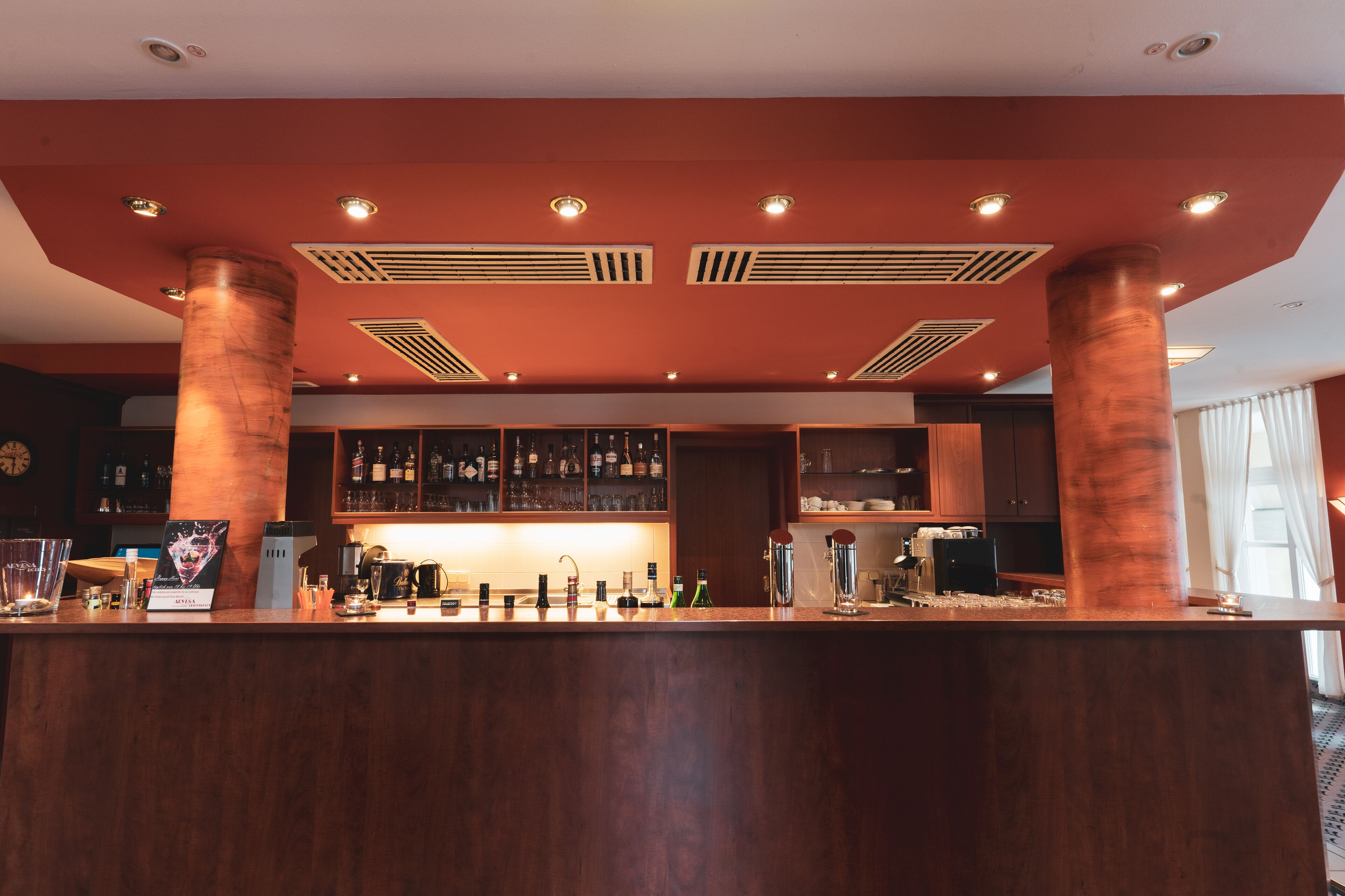 A bar area with a wooden counter, shelves with bottles, and a modern ceiling design.