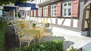 An outdoor dining area with yellow tables and white chairs, set against a building with red shutters and a half-timbered facade.