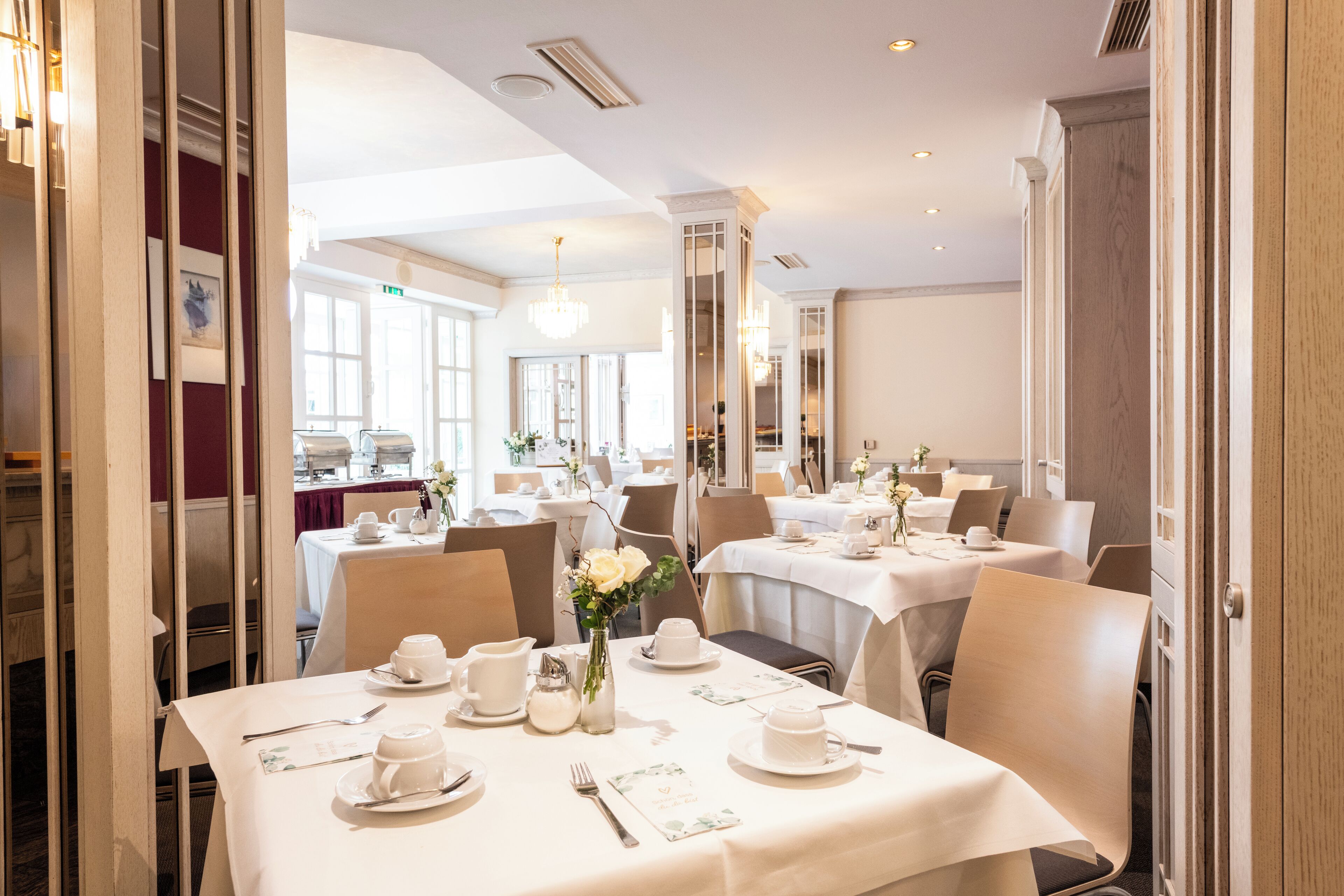 Une salle à manger bien éclairée, avec des tables dressées pour le service, ornées de nappes blanches, d’assiettes et de couverts.