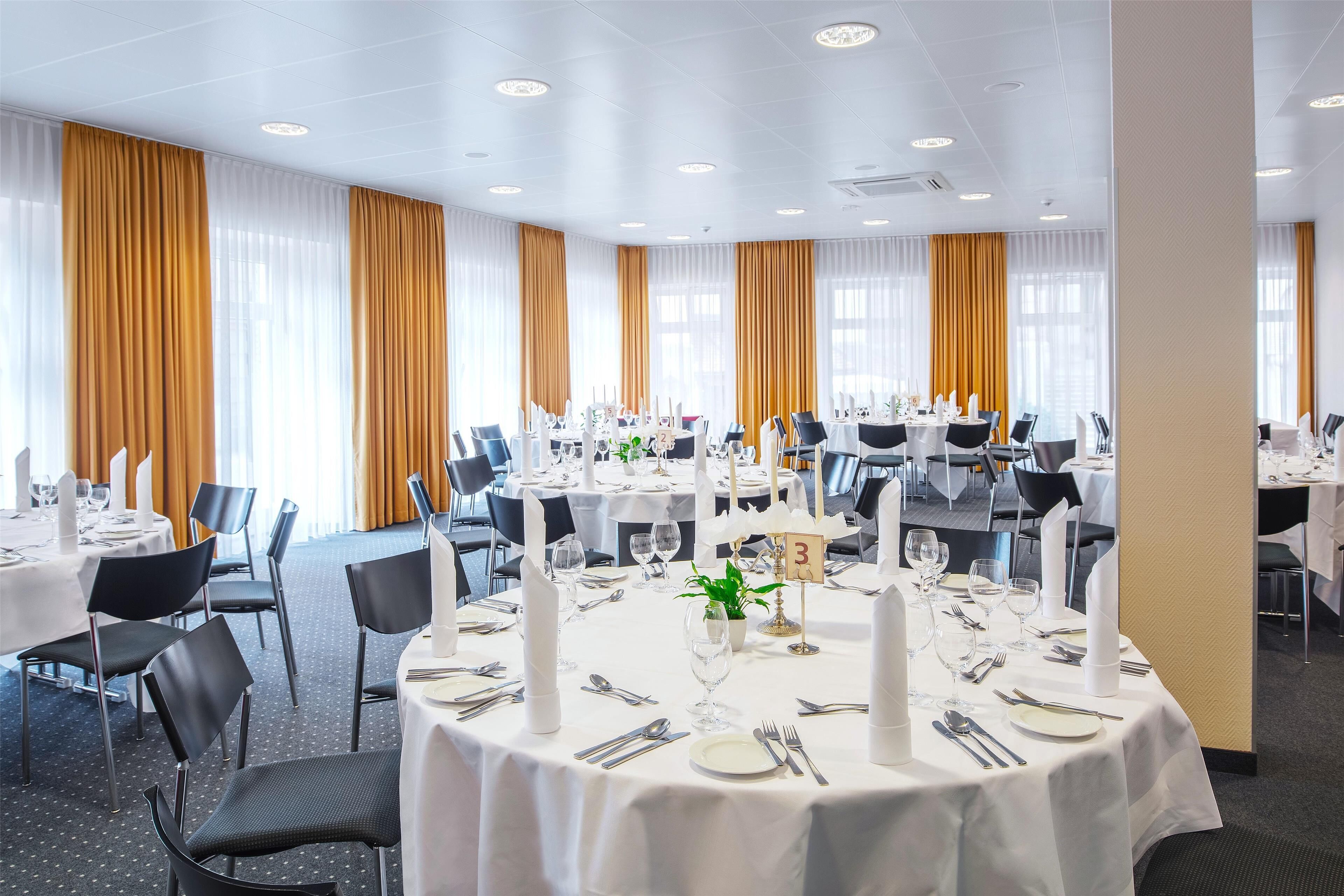 Une salle à manger d’hôtel avec des tables rondes dressées pour le repas, agrémentée de nappes blanches, de chaises noires et de nombreuses bougies.