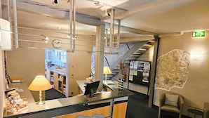 A reception desk with a computer, a lamp, and a bulletin board with information. There is a chair and a table in the corner. A staircase is visible in the background.