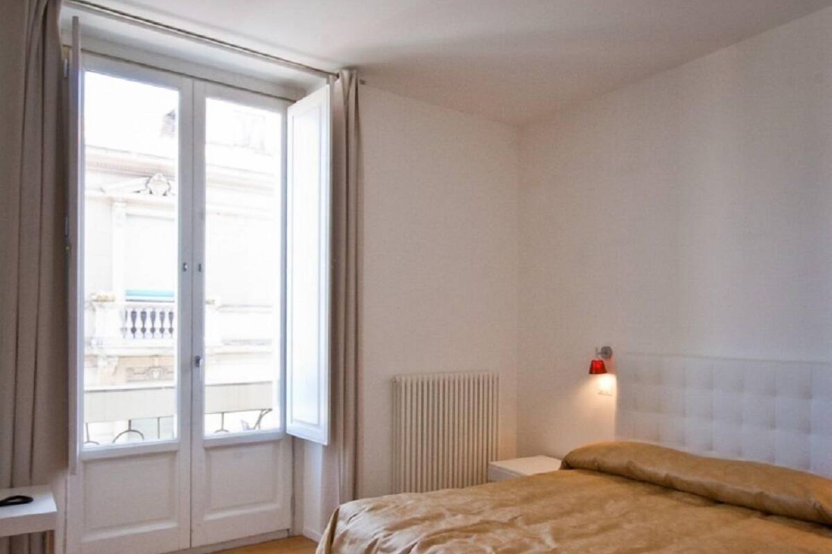 Ein Schlafzimmer mit einem Bett, einem Heizkörper, einem Fenster mit Vorhängen und einem kleinen weißen Tisch.