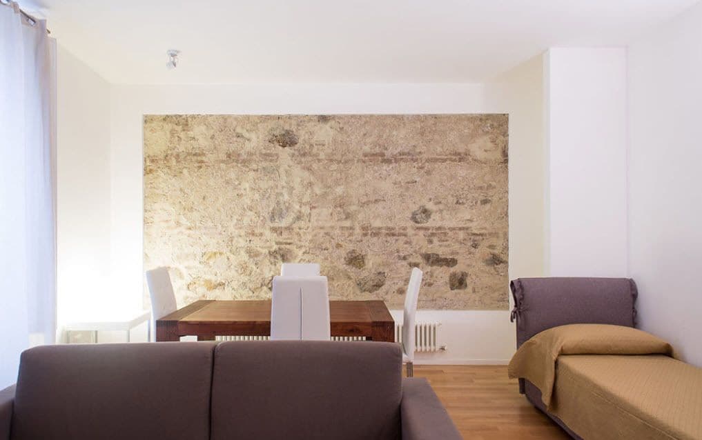 A modern living room with a stone accent wall, a wooden dining table, white chairs, a brown sofa, and a bed with a brown cover.