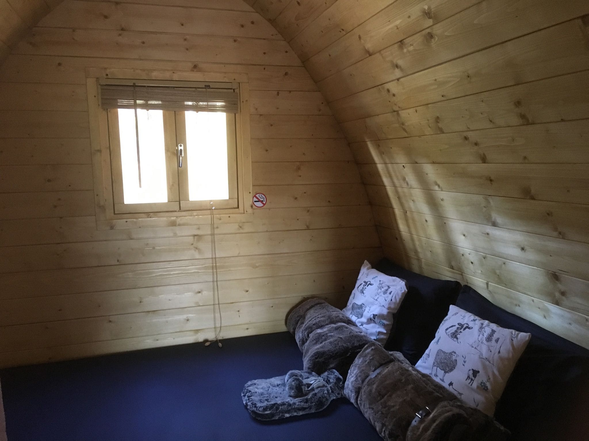 A small, cozy room with wooden walls and a sloped ceiling. There is a window with a metal shutter, a blue mattress on the floor, and two pillows with animal prints.