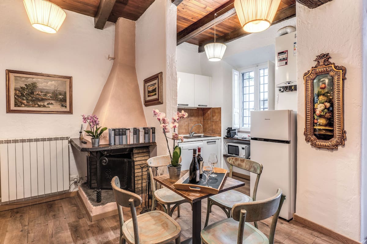 A cozy dining area with a wooden table, chairs, and a fireplace. A kitchen is visible in the background with white cabinets and a refrigerator.